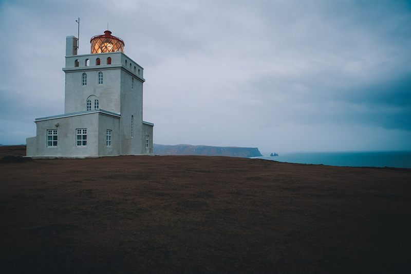 White lighthouse with its lamp lit on a brown headland at dusk, dark cliffs and the sea behind