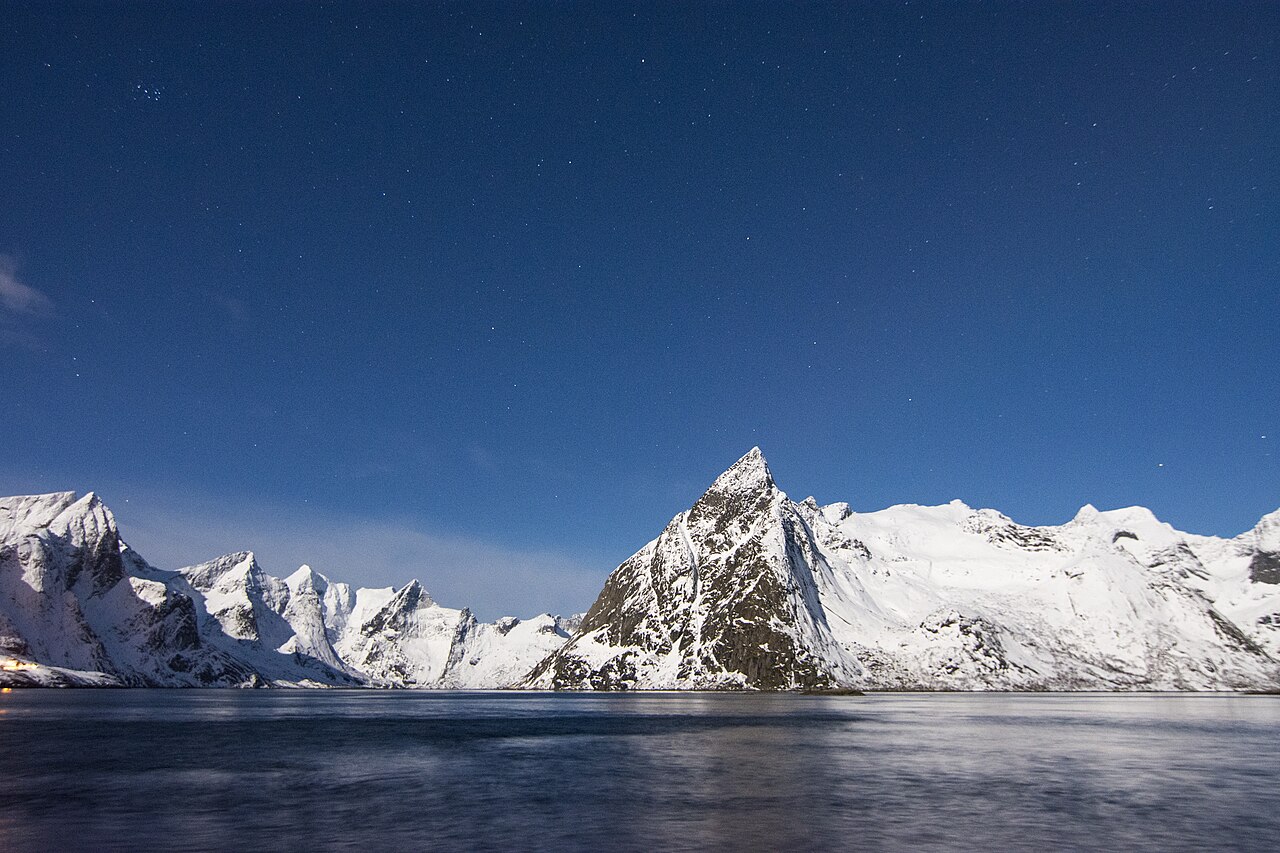 Moonlit winter night over Reinefjorden: snow-covered peaks, calm dark water and a deep blue starry sky