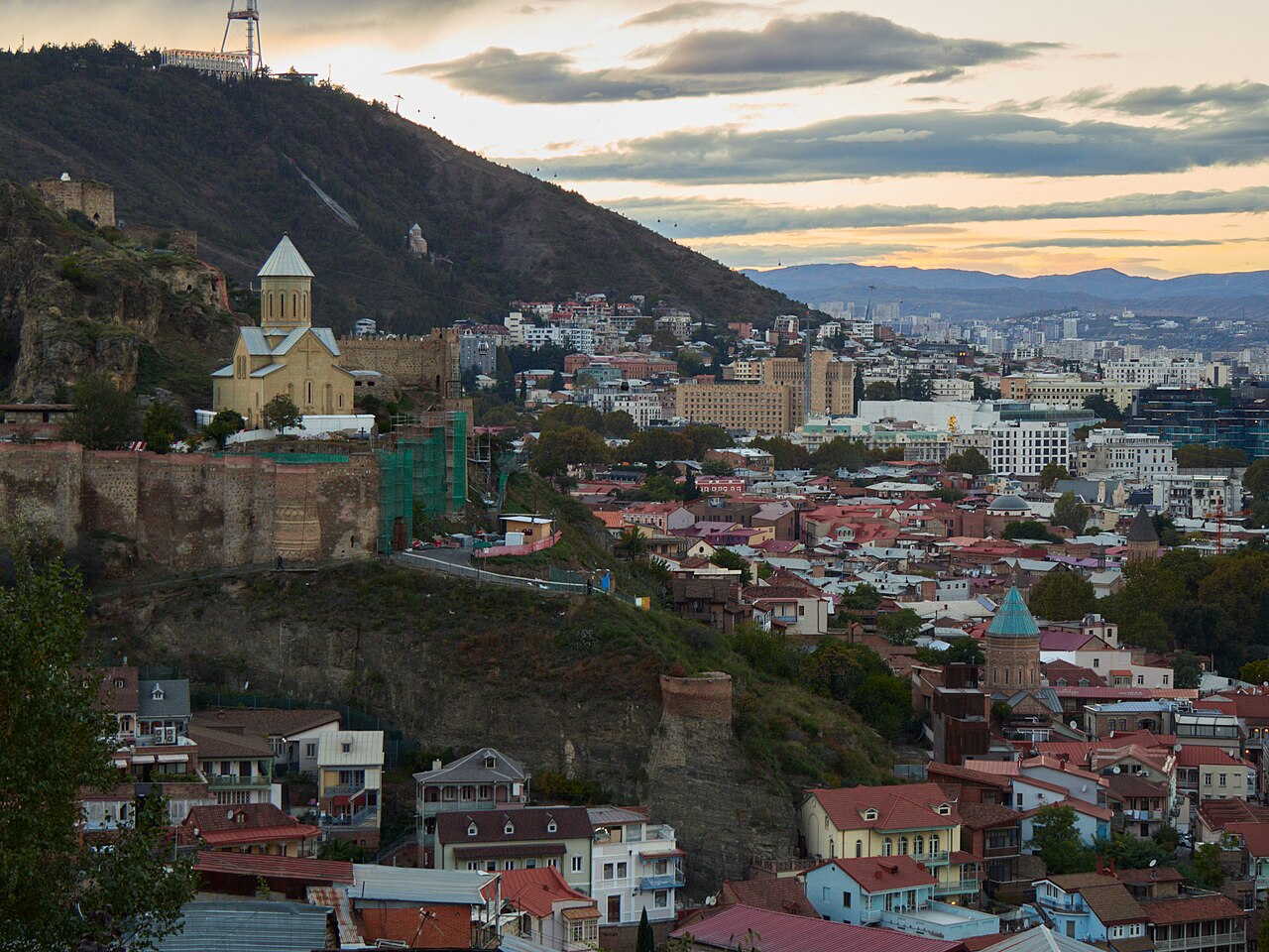 Narikala fortress and the tiled rooftops of old Tbilisi under a soft sunset sky, the city and mountains behind